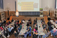 Personen sitzen sich an langen Tischreihen in einem Saal gegenüber