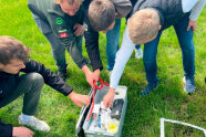 4 Personen beugen sich über Koffer, der auf Wiese steht